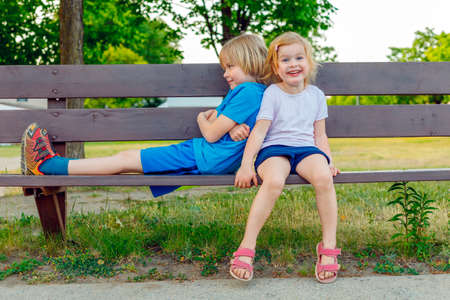 Children sitting on a bench in park in summer. Happy kids having fun. Smiling friends playing outdoors togetherの写真素材
