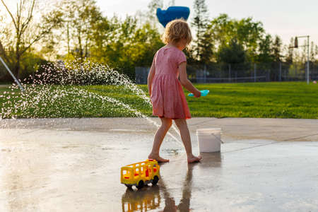Little girl playing with toys at splash pad playground in park in summer. Small child from behind near fountains spending time outdoorsの写真素材