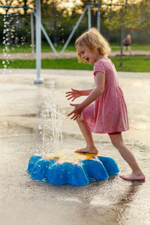 Happy smiling child playing with toys and water in park splash pad. Girl at playground in summer barefoot, enjoying summertimeの写真素材