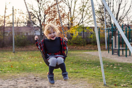 Smiling happy girl on swing in park in autumn. Girl at playground in fall season.の写真素材