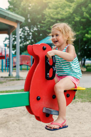 Little girl playing in park at playground . Smiling child sitting on a seesaw swing on a sunny summer day. Happy childhoodの写真素材