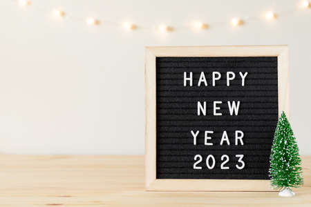 Happy new year 2023 greetings on black felt letter board and small Christmas tree on table and holiday lights on white wall. Copy space for textの写真素材