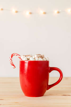 Hot chocolate in red mug with small marshmallows on wooden table with holiday lights on wall. Cocoa in a cup, hot winter drinkの写真素材