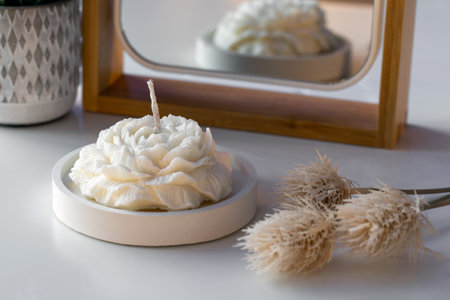White candle in the shape of a flower on the table near the mirror with reflection and lights in the background. Cozy home interior. Spa and candle making concept.の写真素材