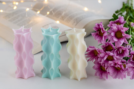 Three soy candles on a table with a book, flowers and lights in background. Home decor. Cozy interior.の写真素材