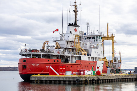 Prescott, Canada - April 6, 2025: red and white coast guard vessel Griffon docked at concrete pier. Ship with canadian flag, heavy cranes, and shipping containers on deck under cloudy sky.のeditorial素材