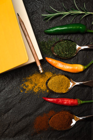 Composition of yellow notebook for recipe and pencil and oriental spices in spoons and peppers and rosemary on the black metallic tray in Asian styleの写真素材