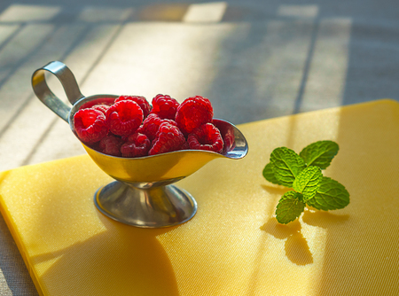 Ripe sweet raspberries in silver bowl and mint on yellow kitchen board in lights of sun. Close up, healthy breakfast ingredient, top viewの写真素材