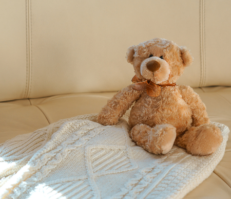 Teddy Bear with ribbon sitting on cozy knitted white Christmas sweater on beige leather sofa background. Warm comfortable winter clothes. Idea for Merry Christmas birthday gift. Copy space, close-upの写真素材