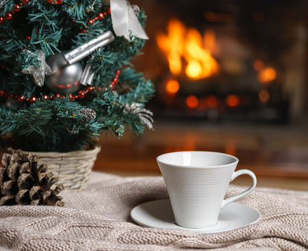 Cup of tea or coffee, woolen knitted things plaid and Christmas decorations near cozy fireplace background, in country house. Cozy relaxed magical atmosphere home interior. New Year winter concept.の写真素材