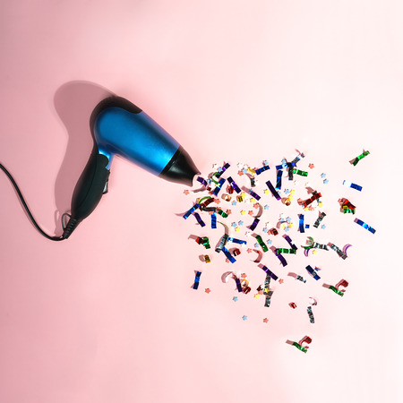 Bright colors confetti flying from blue hair dryer on pastel pink background. Christmas New year winter holiday party decoration. minimalism layout. Top view, copy space, flat layの写真素材