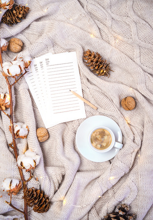 cup of coffee with hearts shapes, pages of diary for writing new plans or shopping gifts, branch of cotton, pine cones and nuts on beige knitted plaid. Cozy winter autumn holiday layout, flat lay.の写真素材
