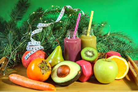 Christmas healthy concept fruits and vegetables. Keto diet food ingredients bottles smoothies acai berry measuring tape dumbbell kiwi avocado pomegranate orange pear fir branches green background.の写真素材