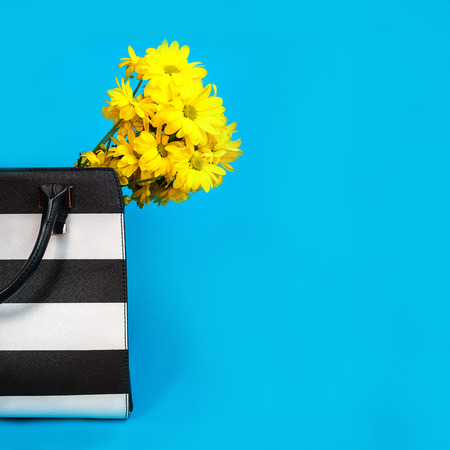 Concept of gift for a business woman: female business accessories black and white bag and yellow flowers on blue background. Valentines Day 8 March birthday party outfit layout, office. Copy spaceの写真素材