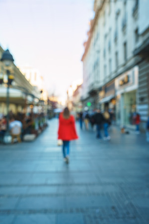 Blurred background of young woman in red coat walking down the street on the center of city with buildings in the background. Sunset time. Defocused background.の写真素材