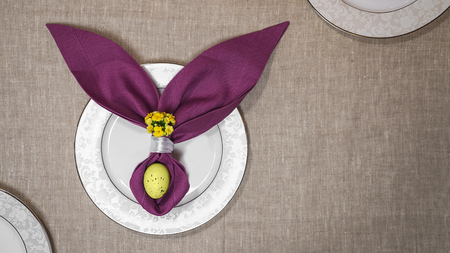 Beautiful Easter table setting with egg, purple napkin Easter Bunny on white plates and on natural tablecloth background. Flat lay, top view.の写真素材