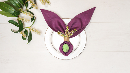 Beautiful Easter table setting with egg, purple napkin Easter Bunny, spring green tree branch on white wooden background. Flat lay, top view. Copy space.の写真素材