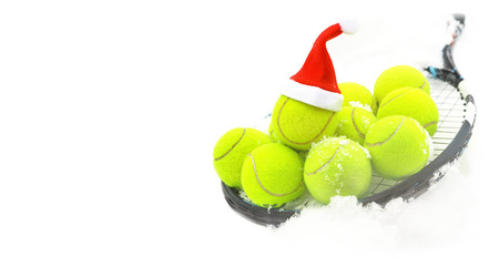 Tennis winter concept with tennis balls and racket on white snow, isolated. Top view, copy space. Christmas New Year sport healthy layout. Santa Claus hat on the top off ball. Banner size.の写真素材