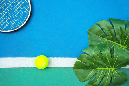 Summer sport concept with green monstera leaf and tennis ball, racquet on white line on hard tennis court. Flat lay, top view, copy space. Blue and green colors.の写真素材
