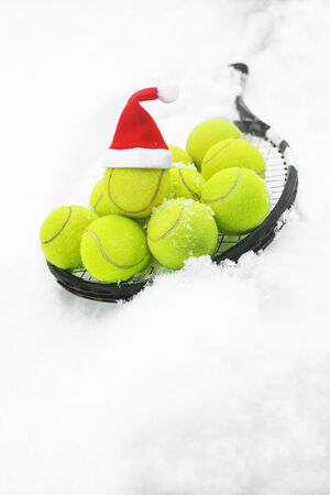 Tennis winter concept with tennis balls and racket on white snow, isolated. Top view, copy space. Merry Christmas and Happy New Year sport healthy layout. Santa Claus hat on the top off ball.の写真素材