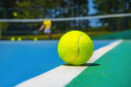 Tennis ball on white court line on hard modern blue green court with player, net, balls, trees on the background. Close-up, selective focus. Sport, tennis play, healthy lifestyle concept.の写真素材