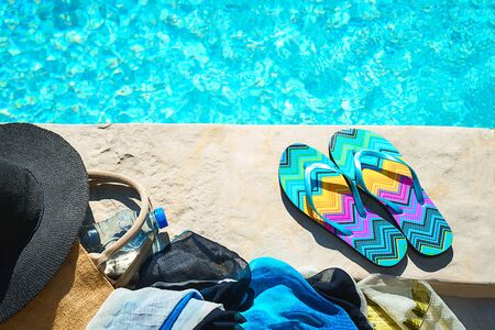 Swimming pool and straw hat, colorful flip flops, wicker bag, straw hat, bottle with still water as sunny summer background. Travel holiday suntan vacation concept. Flat lay, top view, close up.の写真素材