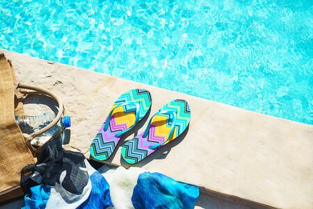 Swimming pool and straw hat, colorful flip flops, wicker bag, straw hat, bottle with still water as sunny summer background. Travel holiday suntan vacation concept. Flat lay, top view, close up.の写真素材