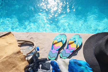 Swimming pool and straw hat, colorful flip flops, wicker bag, straw hat, bottle with still water as sunny summer background. Travel holiday suntan vacation concept. Flat lay, top view, close up.の写真素材