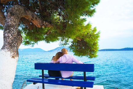 Couple is hugging and sitting close having fun look on the sea on blue bench during summer holiday vacation. Couple make wonderful plans for future, exploring Greece, island Hydra.の写真素材