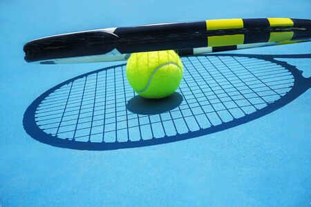 Summer sport concept with tennis ball and racket on blue hard tennis court. Top view, copy space. Blue and yellow, sunlight and shadow.の写真素材