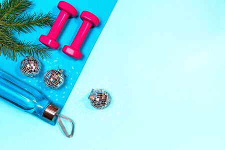 Christmas sport set with pink dumbbells, bottle of water, fir tree branches on blue yoga mat and disco balls decoration on pastel blue background. Flat lay, top view, copy space.の写真素材