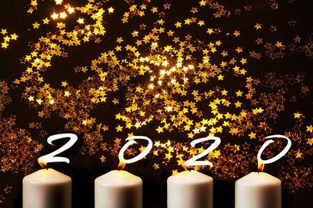 Happy New Year 2020 is written with candle flames on black background with blurred gold stars....