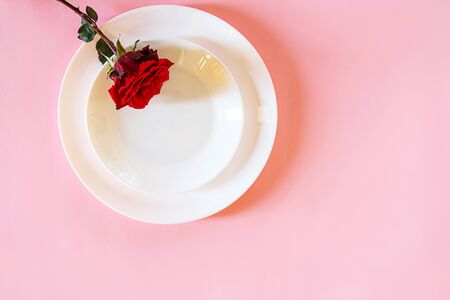 Romantic table setting for Valentines day or dinner date celebration wedding. Valentines day concept with red rose bud white plates on pastel pink tablecloth background. Flat lay, top view copy space.の写真素材