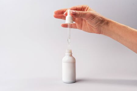 Hand of woman with pipette with drop of serum or hyaluronic acid or collagen moisturizer and white bottle on gray background. Beauty cosmetic consept for hydrating face masks, skin care solutions.の写真素材