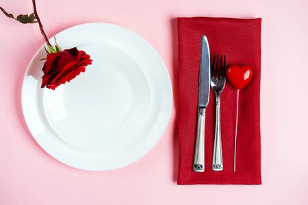 Romantic table setting for Valentines day or dinner date celebration wedding. Valentines day concept with red rose bud white plates on pastel pink tablecloth background. Flat lay, top view copy space.の写真素材