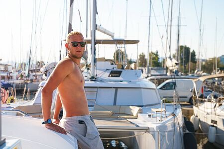 Happy young man feels happy on marina near the luxury sail boat yacht catamaran in summer holidays. Caucasian male model.の写真素材