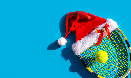 Santa Claus hat on tennis racket and ball on blue background. Merry Christmas and Happy New Year tennis concept. Close up, sport lifestyle, funny.の写真素材