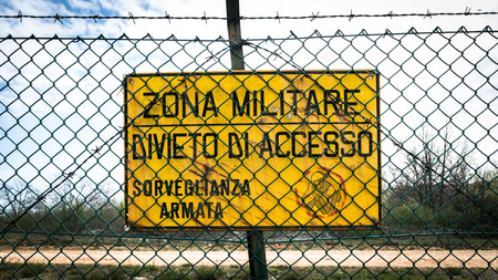 Yellow sign that reads in Italian military zone, no entry, armed surveillance.
