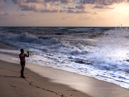 Tropea, Italy - August 26, 2018: Man with smartphone photographs a wave crashing on the beach at sunset.のeditorial素材