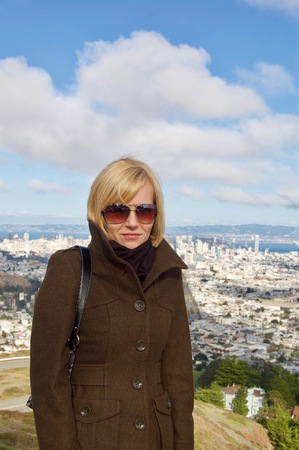 Beautiful blonde female tourist visiting the San Francisco Twin Peaks overlooking the city's scenic skyline with numerous buildings and landmarks in the background on a winter day in California, USAの写真素材