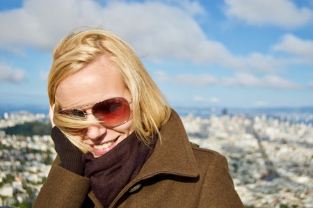 Beautiful smiling blonde female tourist visiting the San Francisco Twin Peaks overlooking the city's scenic skyline with many buildings and landmarks in the background on a winter day in California, USAの写真素材