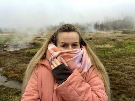 Beautiful blonde caucasian tourist from Europe traveling to The Great Geysir in Iceland in autumn / winter wearing warm clothes (jacket, snow boots) while sightseeing in the surreal landscapeの写真素材