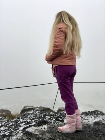 Beautiful blonde tourist girl with long blonde hair and pink winter clothes visiting the scenic Gullfoss Waterfall on the Golden Circle Road in Iceland on a foggy autumn day, standing on black rocksの写真素材