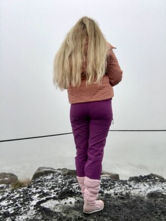 Beautiful blonde tourist girl with long blonde hair and pink winter clothes visiting the scenic Gullfoss Waterfall on the Golden Circle Road in Iceland on a foggy autumn day, standing on black rocksの写真素材