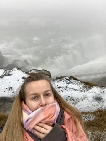 Isolated caucasian woman with long blonde hair on a sightseeing adventure in the iceland landscape at Gullfoss waterfall wearing a salmon pink winter jacket and a scarf to protect from the wind & snowの写真素材