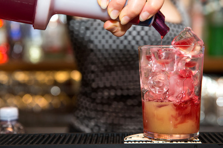 a close up of a barman making a tequila sun rise in a tumbler glass. Barmaid is pouring some red syrup into the glass.の写真素材