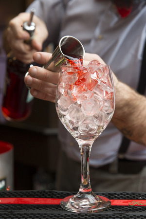 closeup of a barman making a cocktail and pouring red liquid into a wine glass full of ice cubesの写真素材