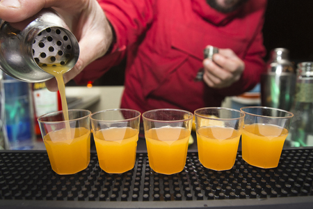 bartender is pouring an alcoholic orange cocktail into small glasses on the counterの写真素材