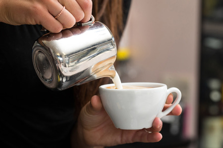 close up of a young woman pouring milk into a cup of coffee, he is making a cappuccinoの写真素材