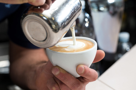 close up of a man pouring milk into a cup of coffee, he is making a cappuccinoの写真素材